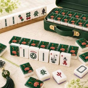 May include: A Mahjong set featuring white tiles with green backs decorated with pink lotus flowers and orange koi fish. The set includes a dark green carrying case, a wooden rack, and tiles displaying various Chinese characters, numbers, and a Joker symbol.