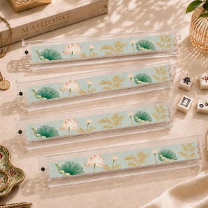 May include: Four clear acrylic racks for holding game tiles, each featuring a light blue fabric insert decorated with a repeating pattern of pink lotus flowers, green lily pads, and gold-toned fern-like foliage, arranged in a vertical stack.