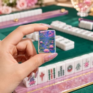 May include: A rectangular mahjong tile featuring a vibrant, colorful illustration of pink lotus flowers floating on a blue pond. The tile is held upright, displaying the detailed floral artwork against a backdrop of other white mahjong tiles.