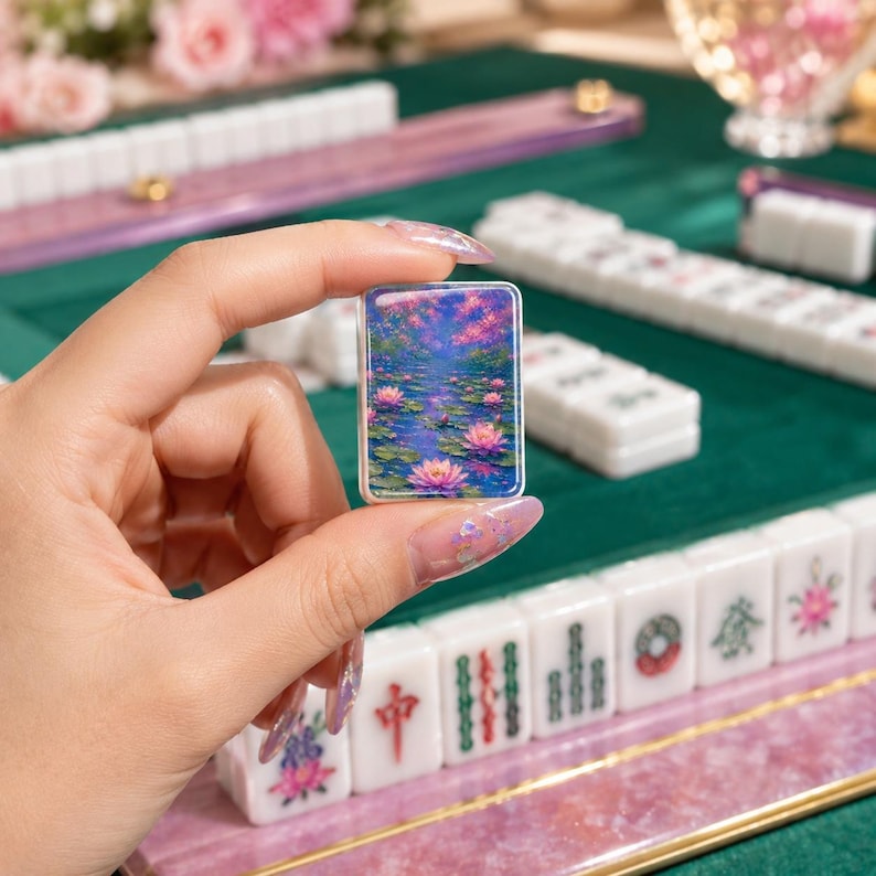 May include: A rectangular mahjong tile featuring a vibrant, colorful illustration of pink lotus flowers floating on a blue pond. The tile is held upright, displaying the detailed floral artwork against a backdrop of other white mahjong tiles.