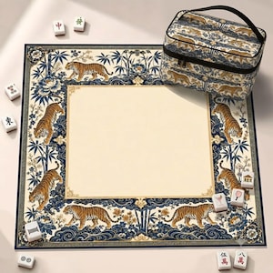 May include: A square mahjong mat featuring a cream-coloured centre bordered by a blue and gold pattern of tigers, bamboo, and flowers. A matching rectangular carrying case with a handle and zip closure sits on the mat, accompanied by scattered white mahjong tiles.