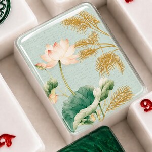 May include: A rectangular mahjong tile featuring a light mint green background with a printed illustration of a pink lotus flower, green lily pads, and gold fern-like foliage. The background includes faint, small, illegible text patterns.