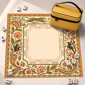 Könnte beinhalten: Eine quadratische Mahjong-Matte mit cremefarbenem Zentrum und aufgedruckten Charleston-Regeln, umgeben von einem orangefarbenen, grünen und floralen Rand mit Affen und einem Leoparden. Enthält eine gelbe Tragetasche und mehrere weiße Mahjong-Steine.