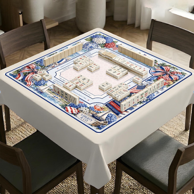 May include: A square table covered with a white tablecloth featuring a nautical-themed border with blue hydrangeas, red and white striped ribbons, sailboats, and the text Liberty & Co. A Mahjong set with tiles is arranged on the table surface.