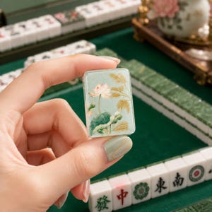 Puede incluir: Una ficha de mahjong rectangular que presenta una ilustraci&oacute;n detallada de una flor de loto rosa con hojas verdes y juncos dorados sobre un fondo verde salvia suave. La ficha tiene un acabado liso y brillante con bordes redondeados.