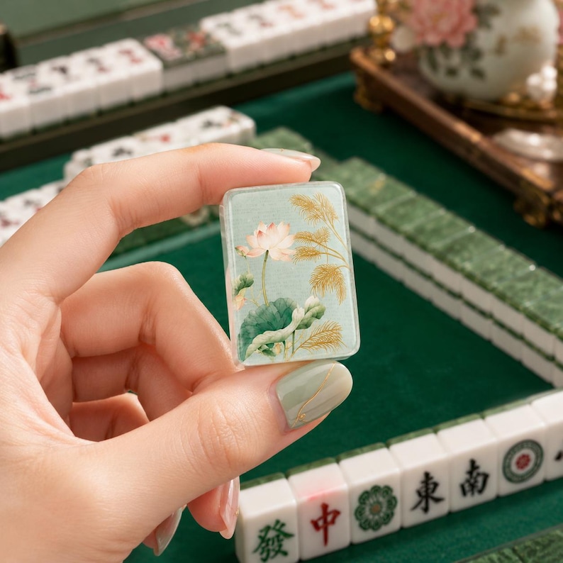 Puede incluir: Una ficha de mahjong rectangular que presenta una ilustraci&oacute;n detallada de una flor de loto rosa con hojas verdes y juncos dorados sobre un fondo verde salvia suave. La ficha tiene un acabado liso y brillante con bordes redondeados.