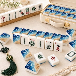May include: A Mahjong set featuring tiles with a blue ocean wave and sandy beach design on the back, alongside traditional tiles with Chinese characters, bamboo, and numbers. Includes a cream-colored carrying case and a green tassel charm.