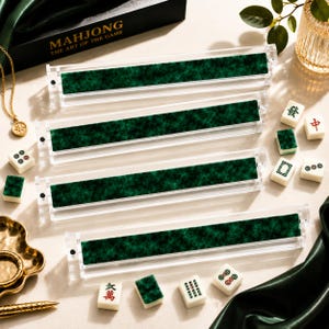 Op de afbeelding: Vier doorzichtige acryl mahjong-tegelhouders met een donkergroene vilten voering. De set wordt vergezeld door verschillende verspreide witte mahjong-tegels met rode, groene en zwarte markeringen, naast een zwarte doos met de tekst MAHJONG THE ART OF THE GAME.