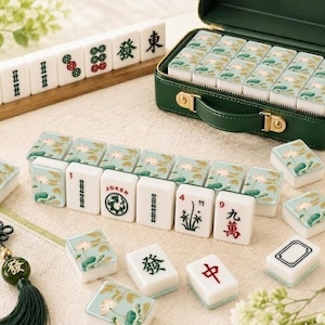 Puede incluir: Un juego de mahjong con fichas blancas que presentan motivos de flores de loto verdes en el reverso y caracteres o s&iacute;mbolos tradicionales en el anverso. El set incluye un malet&iacute;n de transporte verde oscuro y un soporte de madera.