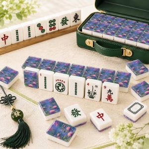 May include: A Mahjong set featuring white tiles with green and red traditional characters, alongside tiles decorated with a blue and pink water lily pond print. The set includes a dark green storage case and a green tassel with a character bead.