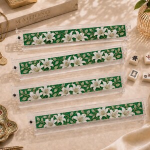Może przedstawiać: Cztery przezroczyste akrylowe stojaki na płytki mahjong z ozdobną wkładką przedstawiającą powtarzający się wz&oacute;r białych kwiat&oacute;w lilii i zielonych liści na ciemnozielonym tle. Każdy stojak posiada małą czarną śrubkę po lewej stronie.