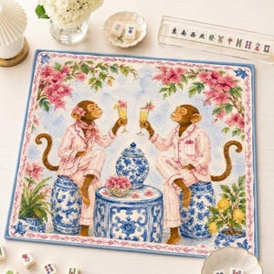 Op de afbeelding: Een vierkante stoffen mat met een aquarelillustratie van twee apen in roze gestreepte pyjama's, zittend op blauw-witte keramische tuinkrukjes, proostend met gele drankjes, omringd door roze bloemen en een blauw-witte pot.