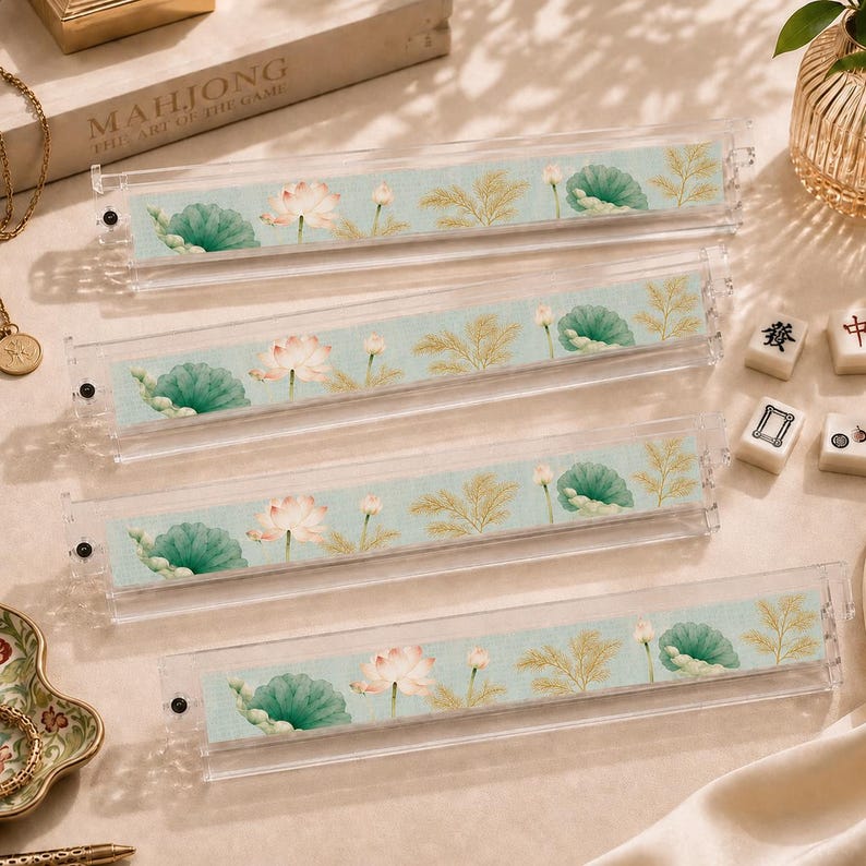 Puede incluir: Cuatro soportes de acr&iacute;lico transparente para fichas de juego, cada uno con una inserci&oacute;n de tela azul claro decorada con un patr&oacute;n repetitivo de flores de loto rosas, nen&uacute;fares verdes y follaje dorado, dispuestos en una pila vertical.