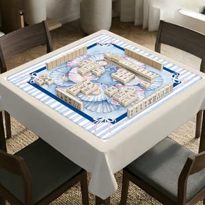 May include: A square mahjong table cover featuring a central watercolor illustration of blue and white seashells with pink ribbons. The border has a dark blue frame and light blue and white stripes. Mahjong tiles are arranged on the surface.