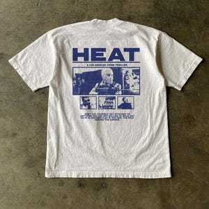 May include: A white short-sleeve t-shirt featuring a blue graphic on the back. The design includes the text HEAT A LOS ANGELES CRIME THRILLER, a central image of a masked figure, two smaller scene panels, and a quote about walking out in 30 seconds.