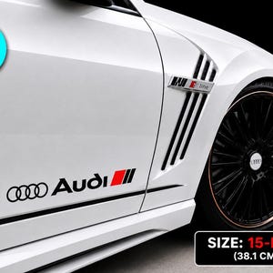Può includere: Un set di due adesivi in vinile nero da 38,1 cm per auto bianca, con il logo e la scritta Audi accompagnati da una striscia decorativa rossa e nera. L'adesivo è applicato sul pannello laterale inferiore, vicino alla ruota anteriore.