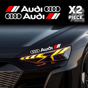 May include: A set of two white Audi vinyl decals featuring the brand logo and a red and white striped graphic. The stickers are displayed on the bonnet of a dark grey car, positioned above the illuminated yellow headlight assembly.