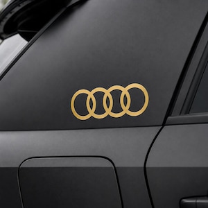 Op de afbeelding: Een goudkleurige vinylsticker met het logo van de vier in elkaar grijpende ringen van Audi, aangebracht op het matzwarte buitenpaneel van een voertuig. De sticker bestaat uit vier dunne, cirkelvormige lijnen in een horizontale rij.