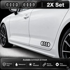 May include: A 2X set of black Audi four-ring logo vinyl decals applied to the lower side panel of a white car. The text 2X Set is visible at the top, and the text Available in 6 Inch & 9 Inch is at the bottom with icons for vinyl, waterproof, UV resistant, adhesive, and installation.