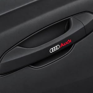 Può includere: Una maniglia per portiera auto nero opaco con il logo Audi in bianco e la scritta Audi in rosso. La maniglia è integrata in un pannello portiera nero opaco abbinato, caratterizzato da un design elegante e sagomato con una finitura uniforme.