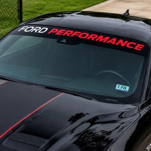 Puede incluir: Un coche negro con una franja en el parabrisas que dice Ford Performance en letras blancas y rojas. El vehículo tiene un acabado brillante, una franja de carreras roja en el capó y una pequeña pegatina de registro en el parabrisas.