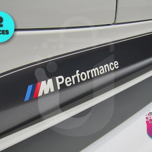 Op de afbeelding: Een wit zijpaneel van een auto met een zwarte horizontale sticker met de tekst M Performance in witte letters. Het logo bevat een schuin blauw, paars en rood streepontwerp. Een cirkelvormige cyaan badge linksboven toont de tekst 2 PIECES.