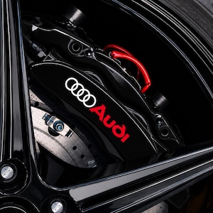 Può includere: Una pinza freno nera ad alte prestazioni con il logo Audi bianco e la scritta Audi rossa. Il componente è montato all'interno di un gruppo ruota nero lucido, mostrando un disco freno metallico con fori di ventilazione visibili e dettagli.
