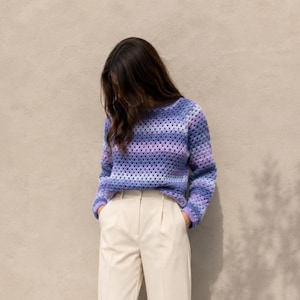 May include: A long-sleeved crochet sweater featuring a granny square pattern in a gradient of purple, lavender, and blue hues. The garment has a round neckline and a relaxed fit, paired with solid cream-colored trousers with a front pleat detail.