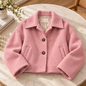 May include: A light pink cropped jacket with a wide collar, long sleeves, and two dark brown buttons down the front. The jacket features two vertical welt pockets on the front and a smooth interior lining visible at the neckline.