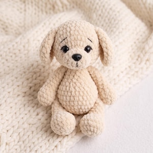 Può includere: Un peluche a forma di cane color crema realizzato in filato morbido e testurizzato. Il pupazzo presenta orecchie pendenti, occhi rotondi scuri, un piccolo naso nero e sopracciglia ricamate, con un motivo a uncinetto visibile sul corpo.