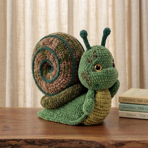 Puede incluir: Un caracol de peluche tejido a ganchillo con una gran concha en espiral en tonos variados de marrón y verde. El caracol tiene un cuerpo verde texturizado, dos antenas erguidas y grandes ojos marrones brillantes con un centro claro.