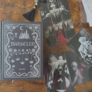 Op de afbeelding: Een zwart gebonden boek getiteld Manacled door Senlinyu met witte lijntekeningen van een kasteel, slangen, rozen en origami-kraanvogels op de kaft. Naast het boek liggen verschillende zwart-wit kunstprints met figuren en een rode mantel.