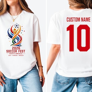 May include: A white t-shirt featuring a colorful graphic on the front with a soccer ball, abstract shapes, and the text 2026 SOCCER FEST NORTH AMERICA USA CANADA MEXICO. The back displays the text CUSTOM NAME above the large red number 10.
