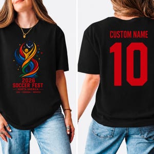May include: A black t-shirt featuring a colorful graphic on the front with a stylized figure, the text 2026 SOCCER FEST NORTH AMERICA USA CANADA MEXICO, and the back displaying the text CUSTOM NAME above the large red number 10.