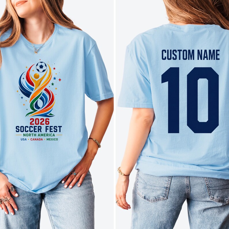 May include: A light blue t-shirt featuring a colorful graphic on the front with a soccer ball, abstract shapes, and the text 2026 SOCCER FEST NORTH AMERICA USA CANADA MEXICO. The back displays the text CUSTOM NAME above the large dark blue number 10.