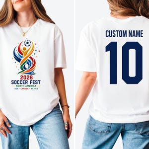 May include: A white t-shirt featuring a colorful graphic on the front with a soccer ball, abstract shapes, and the text 2026 SOCCER FEST NORTH AMERICA USA CANADA MEXICO. The back displays the text CUSTOM NAME above the large dark blue number 10.