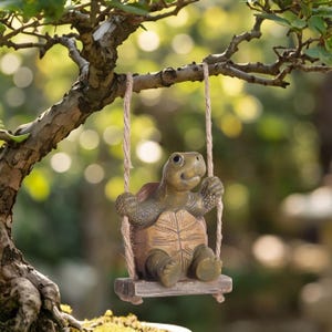 May include: A brown and tan turtle figurine sitting on a small rectangular wooden swing. The swing is suspended by two thick, light brown jute ropes that are tied to a tree branch. The turtle has a textured shell and a smiling facial expression.