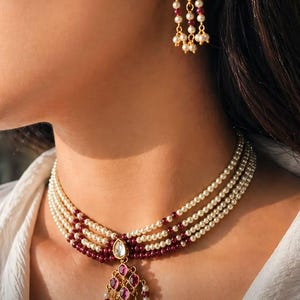 Op de afbeelding: Een meerrijige choker-ketting met rijen kleine witte parels en donkerrode kralen, in het midden voorzien van een goudkleurige hanger met een druppelvormige steen en bungelende parel- en rode kraalaccenten, inclusief bijpassende oorbellen.