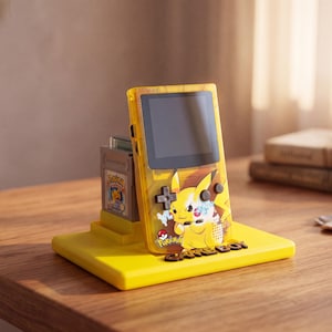 Op de afbeelding: Een gele, doorschijnende handheld spelcomputer met een Pikachu-afbeelding en de tekst Pokémon op de voorzijde. Het apparaat staat op een bijpassende gele, rechthoekige standaard, met een grijze spelcassette in de gleuf aan de achterkant.