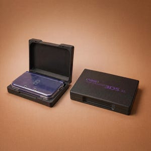 May include: Two black rectangular carrying cases for a handheld gaming console. One case is open, revealing a dark purple console inside, while the other case is closed with the text "new NINTENDO 3DS XL" printed in purple on the top lid.