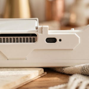 May include: A matte off-white electronic device dock featuring a horizontal ventilation grille with a metallic mesh interior, a centered USB-C charging port, and a small circular indicator light, all integrated into a structured, angular plastic frame.