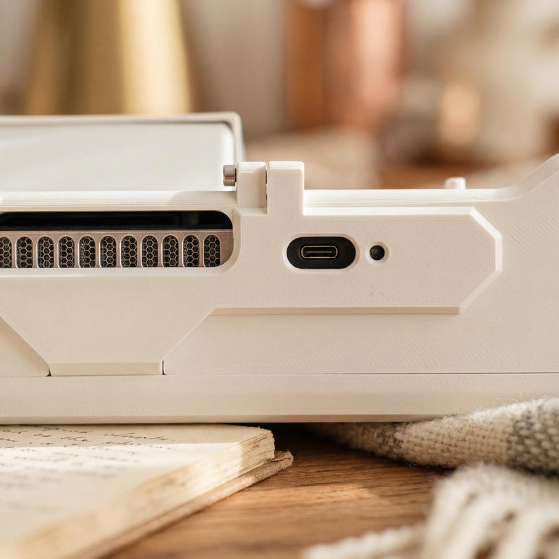 May include: A matte off-white electronic device dock featuring a horizontal ventilation grille with a metallic mesh interior, a centered USB-C charging port, and a small circular indicator light, all integrated into a structured, angular plastic frame.