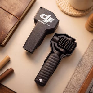 以下が含まれることがあります： 黒いDJI Osmo Pocket 4Pハンドヘルドカメラジンバル。斜めのテクスチャグリップと、DJIロゴおよびOsmo Pocket 4Pの文字が入った保護カバーが付いています。このデバイスは、上部にレンズを搭載したコンパクトで長方形のデザインです。