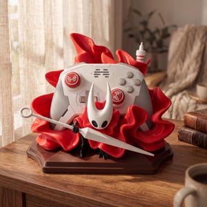 Puede incluir: Un soporte para mando de videojuegos en blanco y gris con una máscara blanca esculpida del personaje Hornet de Hollow Knight, rodeado de elementos rojos vibrantes que parecen tela fluida y una larga aguja blanca sobre la base frontal.