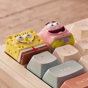 Puede incluir: Dos teclas personalizadas para teclado mecánico con forma de Bob Esponja y Patricio Estrella. La tecla amarilla de Bob Esponja presenta una textura porosa y una cara sonriente, mientras que la tecla rosa de Patricio muestra una amplia sonrisa.