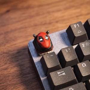 May include: A custom mechanical keyboard keycap shaped like the red and black masked head of the character Deadpool, positioned on a keyboard next to black keys labeled with F1, 1!, 2@, and the Q symbol on a wooden surface.