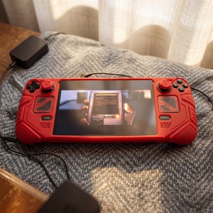 May include: A handheld gaming console encased in a bright red protective silicone cover with textured grips. The device features a black screen displaying a digital interface with text, black joysticks, and standard gaming buttons on the front.