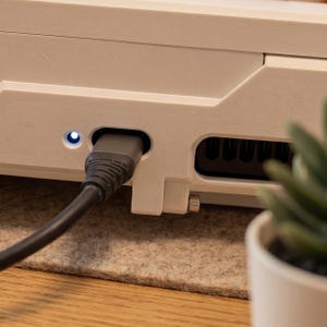 May include: A matte off-white electronic device housing with a textured surface, featuring a black power cable plugged into a port next to a small, glowing circular white LED indicator light and a rectangular ventilation grille with black slats.
