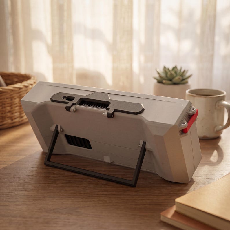 May include: A light gray rectangular electronic device with a black kickstand, a black top-mounted bracket, and a small red accent piece on the side. The device features a ventilation grille on the back and is positioned at an angle on a surface.