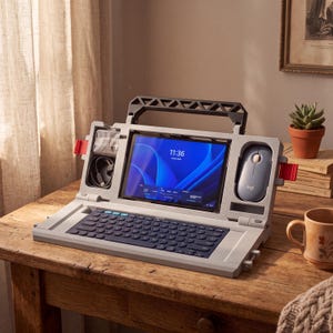 Puede incluir: Una estación de trabajo portátil de color gris claro con soporte para tableta integrado, teclado azul oscuro y compartimentos de almacenamiento para un ratón y cables. La pantalla de la tableta muestra la hora 11:36 y un fondo azul abstracto.